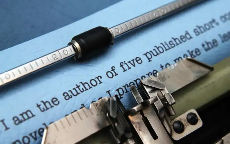 Close-up of a typewriter typing on blue paper, with text reading, "I am the author of five published short..." visible beneath the typewriter's bar and metal type arms.